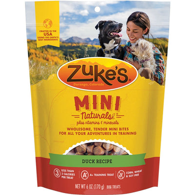 Zuke's Mini Naturals Duck Recipe dog treats in a yellow and red pouch with a woman and two dogs in a mountain landscape.