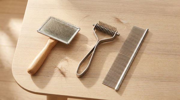 A slicker brush, undercoat rake, and stainless steel dog comb laid out side by side on a light wooden surface