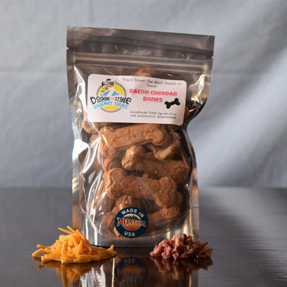 Bacon Cheddar Bones dog treats in clear pouch with shredded cheddar and bacon bits nearby on reflective surface