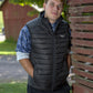 Man leans against rustic red wood fence wearing a black TuffRider Men's Zima Vest outdoors in a rural setting