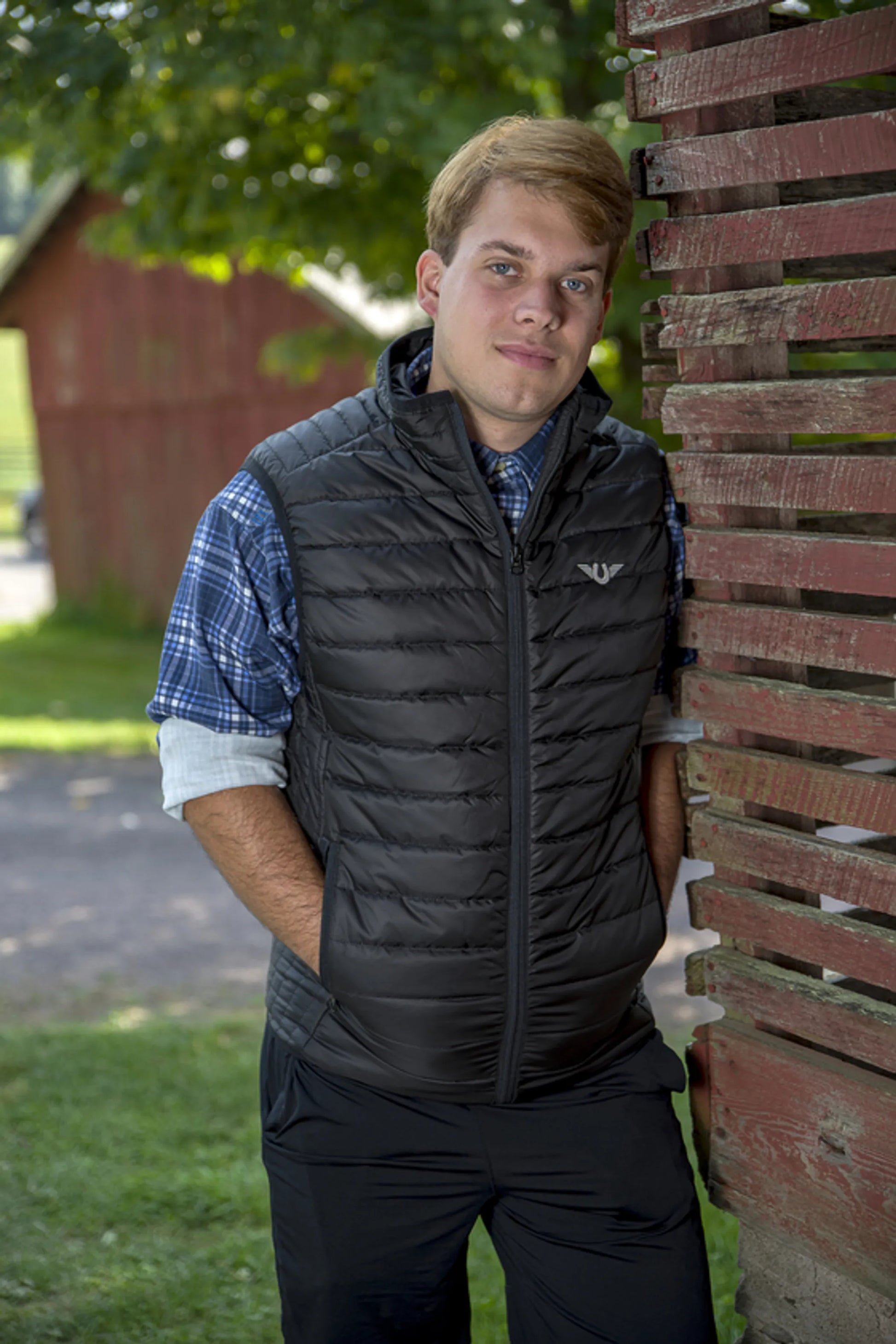 Man wearing black TuffRider Men's Zima Vest standing outdoors next to weathered wooden structure in rural setting