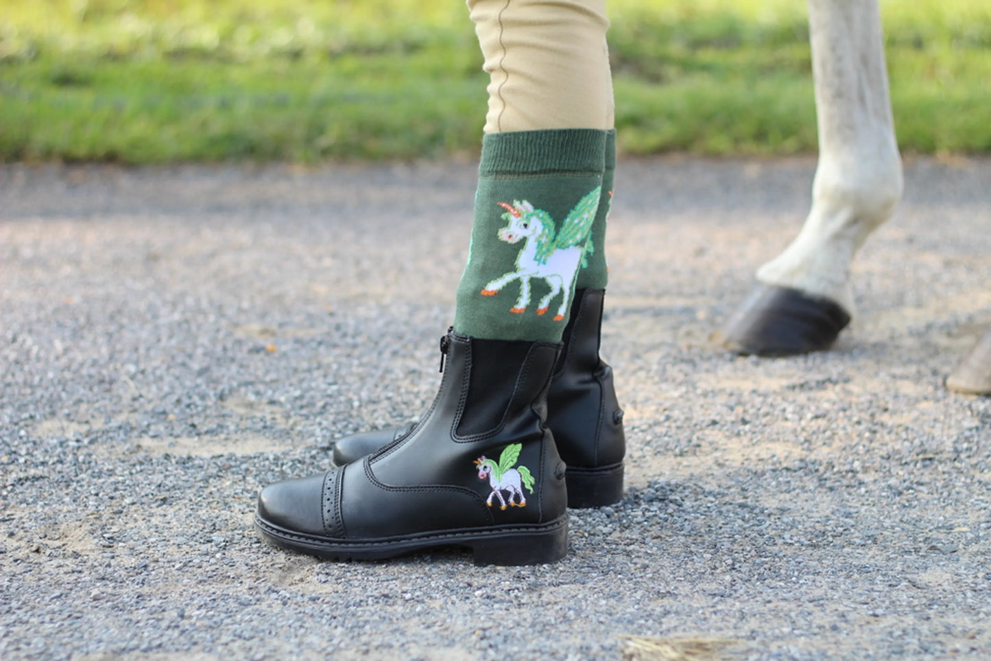 TuffRider Children's Unicorn Starter Front Zip Paddock Boots shown worn with unicorn socks near a horse's hoof.