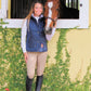 Woman wearing Women's Quilted Classic Vest - Wind-Blocking Equestrian Vest with Snap Front & Dual Zipper - 5/A Baker standing by a horse stable